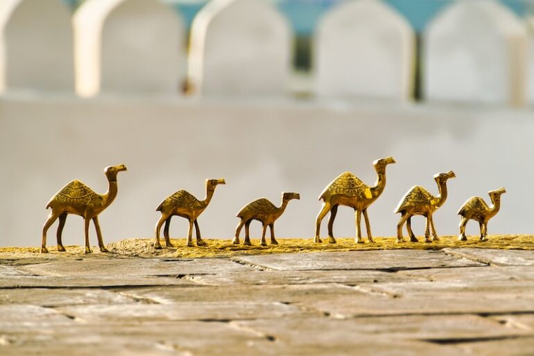 A line of six golden camel figurines of varying sizes, arranged from largest to smallest, on a textured surface with a blurred, light-colored arched structure in the background.
